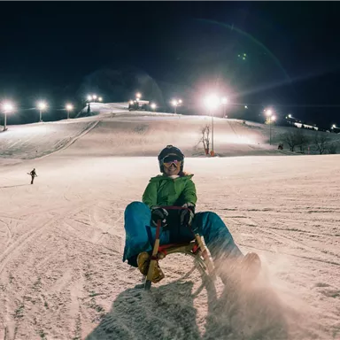 A nighttime sledding fun on a snowy slope. In the background, the lights of the ski slope shine.
