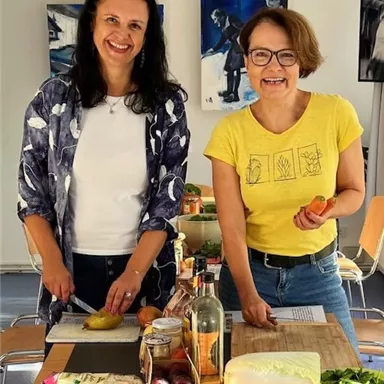 Two women are standing at a table preparing fresh vegetables. The table is covered with various food items and cooking utensils.