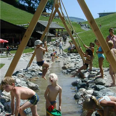 A playground with children playing in the water and on stones. Trees and a building are visible in the background.