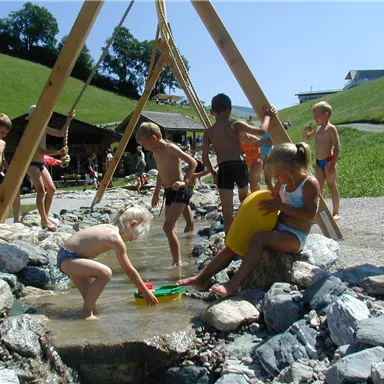 Children are playing in an outdoor water area. There are hills and wooden structures designed for the playground.