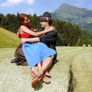 A couple is sitting on a hay bale in a picturesque landscape. In the background, there are green hills and a blue sky.