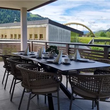 A cozy terrace with a large table and chairs. In the background, there are green hills and a clear blue sky.