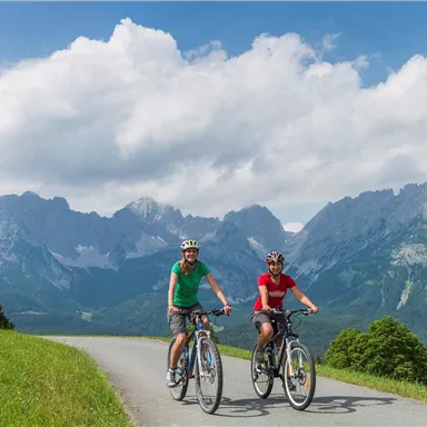 Two cyclists are riding on a path in the mountains. The landscape is green with majestic mountain peaks in the background and a clear sky.