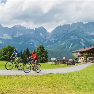 Two cyclists ride on a picturesque path in the mountains. In the background, majestic mountain peaks and a traditional building can be seen.