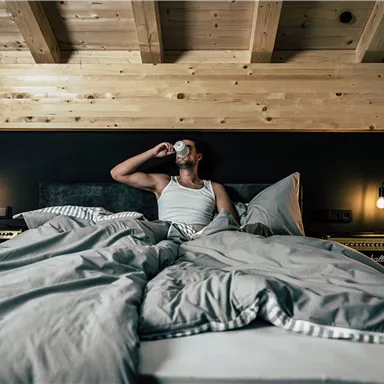A man is sitting in bed and drinking from a cup. The room has a modern decor with a wood-burning stove and a cozy atmosphere.