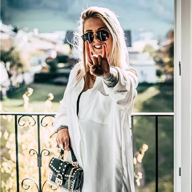 A smiling woman with sunglasses stands on a balcony and displays the peace sign. Behind her, a green landscape with trees and hills can be seen.