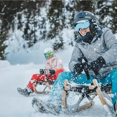 Two people are sledding through the snow. They are wearing winter clothing and helmets and are enjoying the snowy landscape.