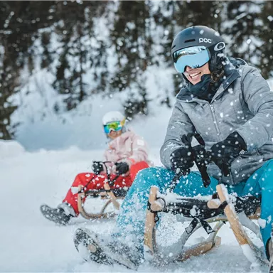 Two people are riding on a sled in the snow. They are having fun and the surroundings are surrounded by trees.