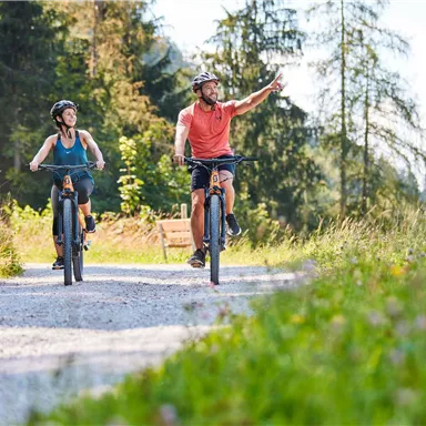 Two cyclists are enjoying a ride on a sunny forest path. A man is enthusiastically pointing in one direction, while a woman is smiling.