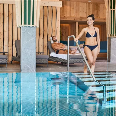 A modern wellness area with a clear pool. A woman in a bikini steps into the water while a man relaxes in the background.