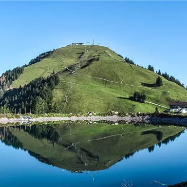 Ein schöner Berg mit grünen Wiesen und Pinienbäumen. Der klare Himmel spiegelt sich im ruhigen Wasser eines Teiches.
