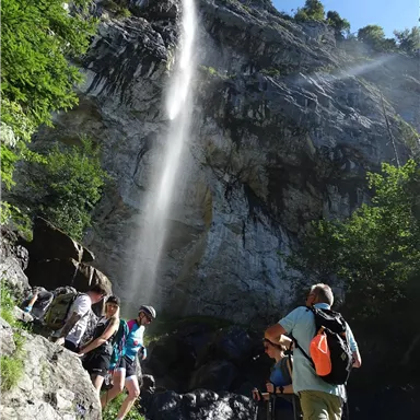 A group of hikers stands before an impressive waterfall in a green, rocky environment. The sky is blue and the sun is shining.