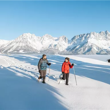 Two people are hiking through a snow-covered landscape. In the background, majestic mountains and a blue sky can be seen.