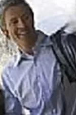 A cheerful man stands on a balcony holding a jacket in his hand. In the background, mountains and trees can be seen.