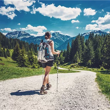 A hiker walks along a gravel path through the mountains. In the background, there are tall mountains and a blue sky with white clouds.