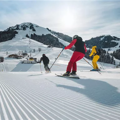 Drei Skifahrer fahren eine präparierte Piste hinunter. Im Hintergrund sind schneebedeckte Berge und ein Skilift zu sehen.