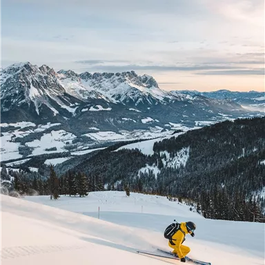 A skier in a yellow suit is skiing down the snowy slope. In the background, majestic mountains and a serene winter landscape can be seen.