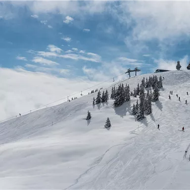 Eine schneebedeckte Berglandschaft mit Skifahrern und Tannenbäumen. Der Himmel ist größtenteils blau mit einigen Wolken.