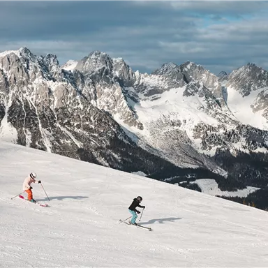Two skiers are descending a snow-covered slope. In the background, impressive mountain ranges are visible.