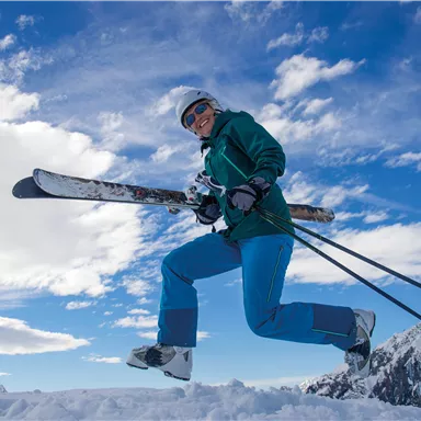 A person jumps over the snow with skis in hand. The sky is clear and the surroundings are mountainous.