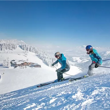 Two skiers are skiing down the slope. The landscape is snow-covered and the sun is shining on a clear blue sky.