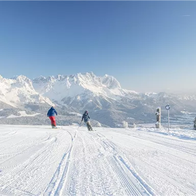 Zwei Skifahrer fahren die verschneite Piste hinunter, umgeben von majestätischen Bergen. Der Himmel ist klar und blau, was einen perfekten Skitag verspricht.