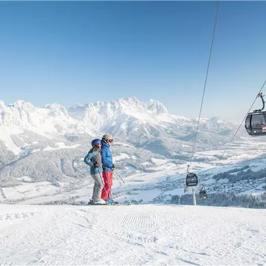 A couple stands on a snow-covered mountain peak overlooking the winter landscape. In the background, the mountains and a cable car can be seen.