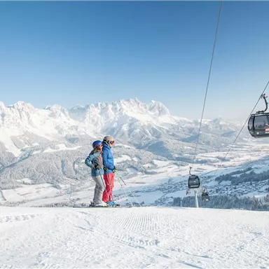 A ski resort with snow-covered mountains and a bright blue sky. Two people are standing on the slope and enjoying the view.