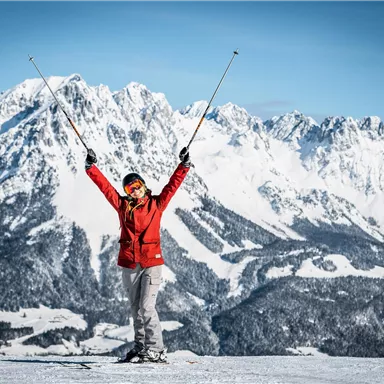 Eine Person steht auf einem schneebedeckten Berg und hebt die Skistöcke in die Luft. Im Hintergrund sind beeindruckende, schneebedeckte Berge zu sehen.