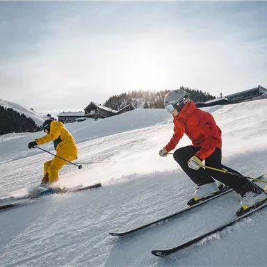 Two skiers are skiing on a snowy slope. The sun is shining and there is snow in the background.