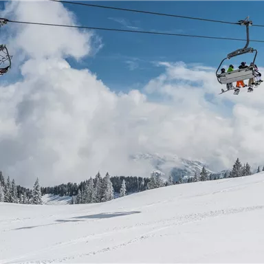 A snowy landscape with a ski lift and trees. The sky is clear and blue with some clouds.