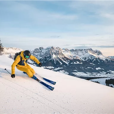 A skier in yellow gear is skiing on fresh snow. In the background, impressive mountains and a clear sky can be seen.
