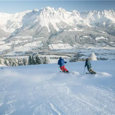 Zwei Skifahrer fahren über eine verschneite Piste. Im Hintergrund erstrecken sich majestätische Berge unter einem klaren Himmel.