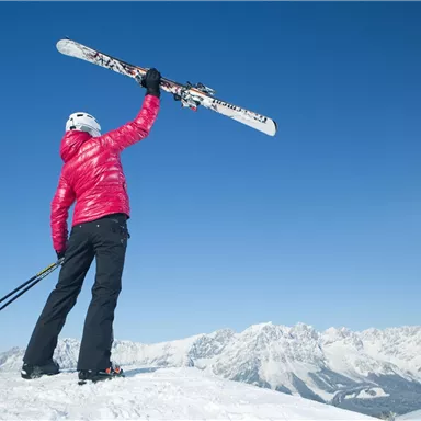 Eine Person steht auf einem schneebedeckten Gipfel und hält ihre Skier in die Höhe. Im Hintergrund sind majestätische Berge und ein klarer blauer Himmel zu sehen.