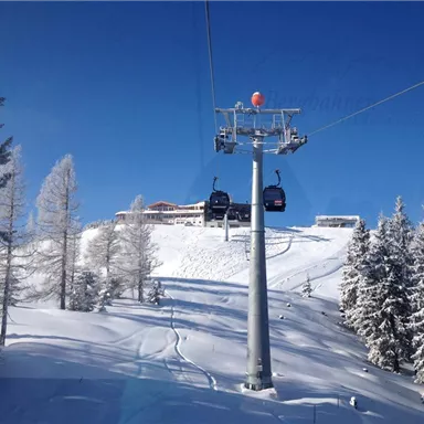 A cable car travels over snow-covered mountains. In the background, snow-covered trees and a building are visible.