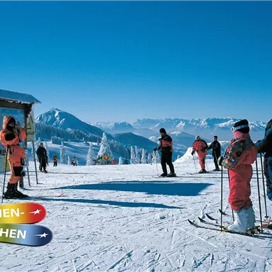 A group of skiers stands on a snow-covered slope with impressive mountains in the background. The scene shows happy families getting ready for ski action.