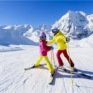 Two children in colorful ski suits are standing on a snow-covered ski slope. In the background, majestic mountains are visible under a clear blue sky.