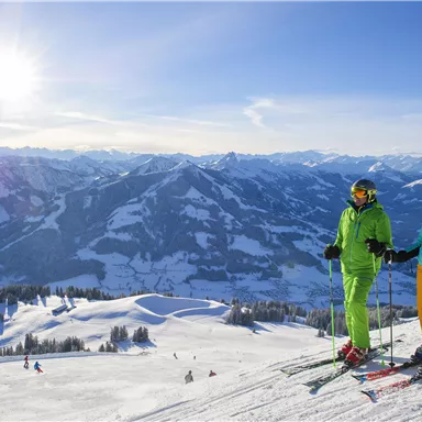 Two skiers stand on a mountain with snow-covered slopes. In the background, majestic mountains and a clear sky can be seen.