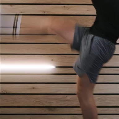 Ein Mann führt einen dynamischen Kick aus. Im Hintergrund sind holzverkleidete Wände und eine moderne Beleuchtung zu sehen.