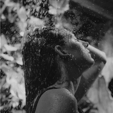 A woman stands under the shower and enjoys the water. The scene is in black and white, with a natural setting in the background.