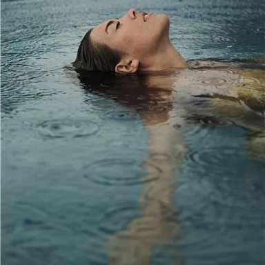 A person lies relaxed in the water while it rains. Their face shows contentment and tranquility.