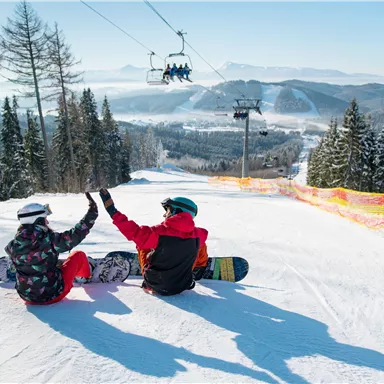 Two snowboarders are sitting in the snow and celebrating together. In the background, snow-covered mountains and a cable car can be seen.