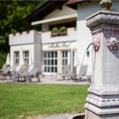 A beautiful fountain stands in the foreground, surrounded by greenery. In the background, there is a building with loungers and umbrellas.