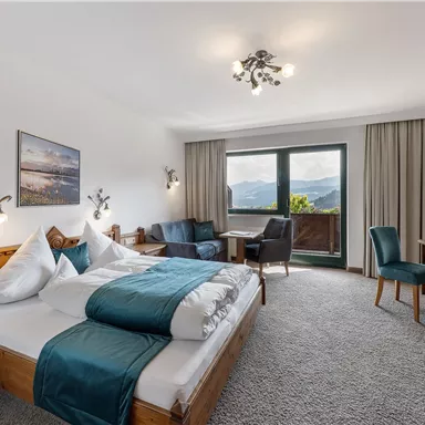 A modern hotel room with a large bed and bright furniture. Through the window, there is a view of the mountains and nature.