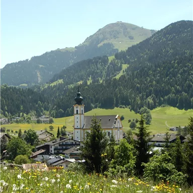 A picturesque landscape with a village and a church in the foreground. In the background, green mountains and a clear blue sky can be seen.