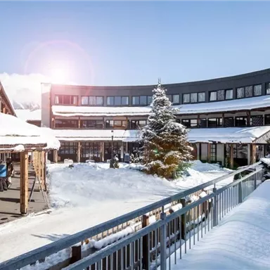 A picturesque courtyard with a snow-covered ground and a Christmas tree. The modern architecture surrounds the space while the sun shines.