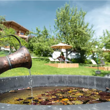 A copper jug is pouring water into a basin filled with leaves. In the background, green meadows and loungers under sunshades can be seen.