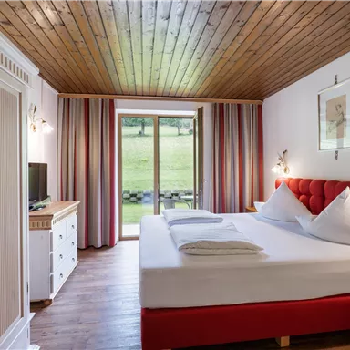 A cozy bedroom with a red bed and wood paneling. There are large windows overlooking greenery, as well as a TV and a chest of drawers.
