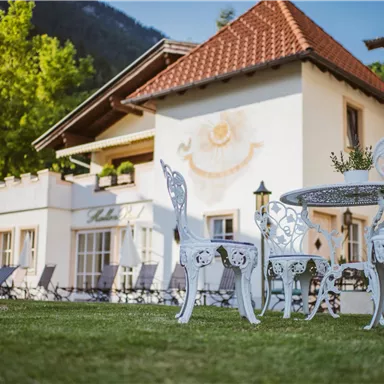 A charming house with a red roof and an inviting garden. In the foreground, elegant white garden furniture sits on green grass.