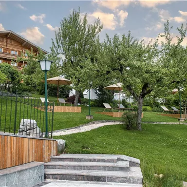 A green garden landscape with several trees and lounge chairs. In the background, there is a beautiful wooden building.